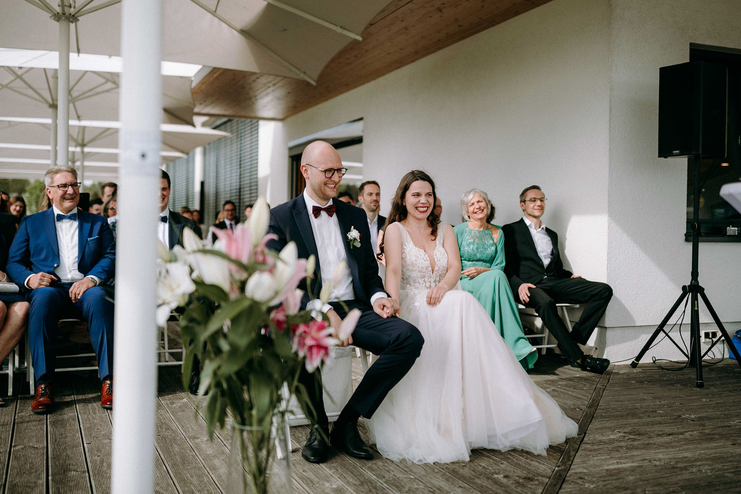 Das Brautpaar lacht herzlich waehrend der Ansprache der Traurednerin auf der Terrasse