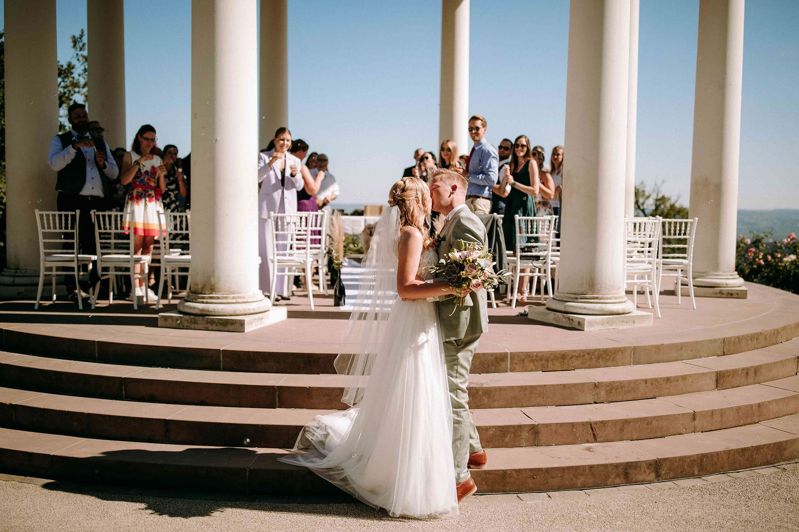 Brautpaar küsst sich vor dem Niederwaldtempel bei ihrer Hochzeit im Rheingau mit applaudierenden Gästen.