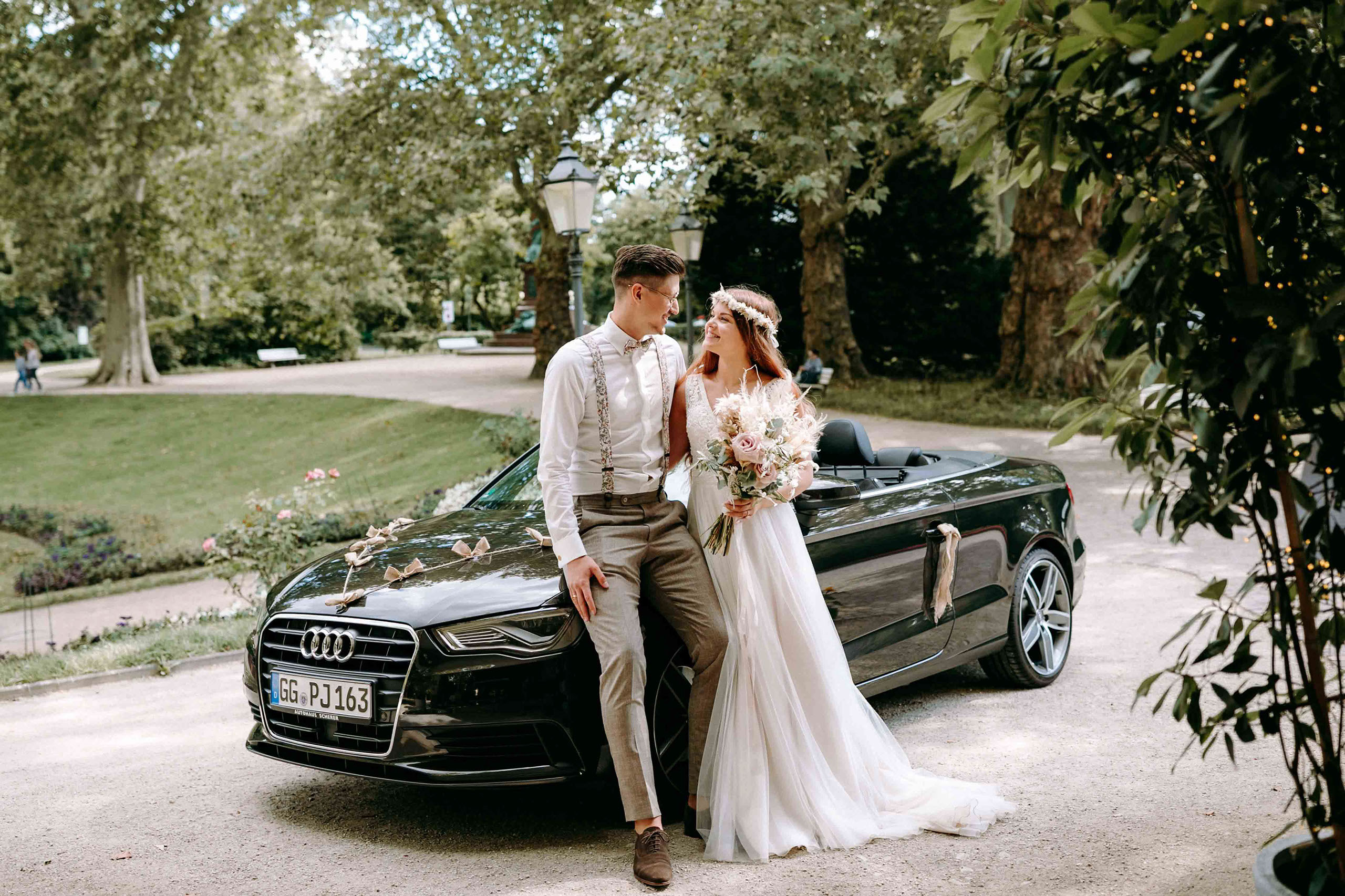 Braut und Bräutigam posieren vor schwarzem Audi Cabrio im Kurpark von Bad Homburg.