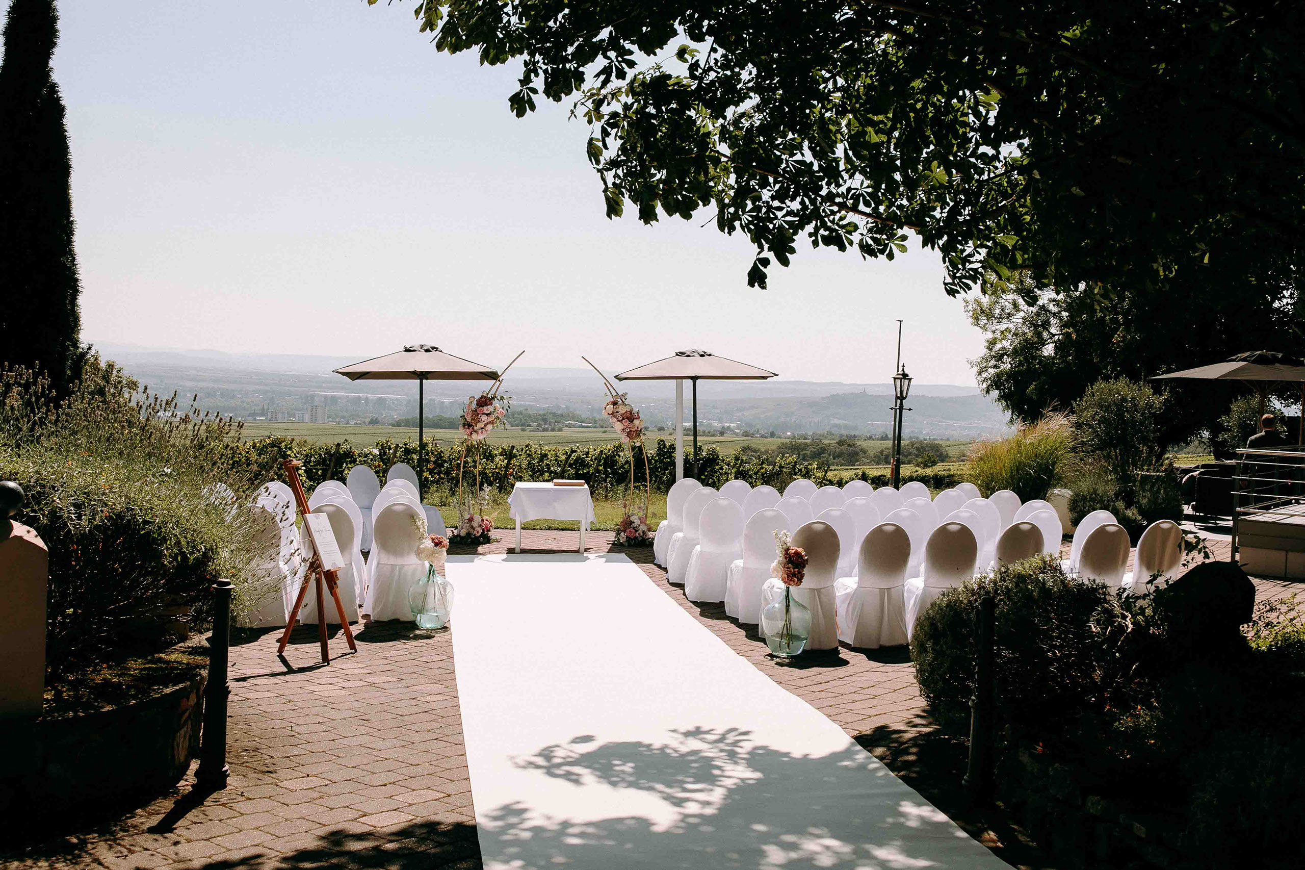 Weiße Stuhlreihen und florale Dekorationen für eine freie Trauung mit Panoramablick auf die Weinberge bei Burg Schwarzenstein – Hochzeitsfotos R