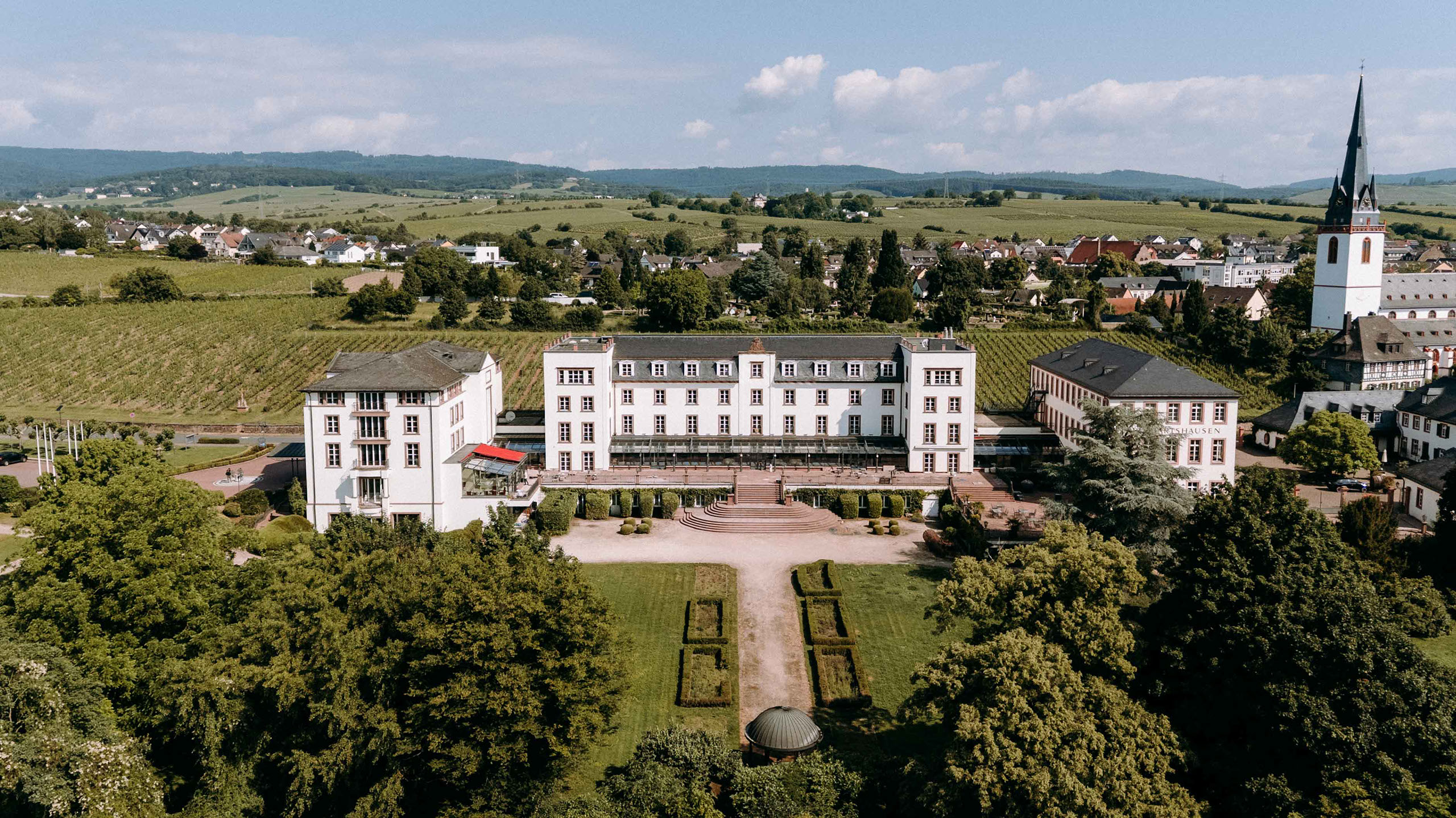 Drohnenbild des Schlosses Reinhartshausen aus leichtem Winkel, mit Blick auf die Weinberge, die historische Kirche und die umliegenden Dächer.